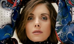 Close-up of the face of Alison Brie, her hands above her head, the puffy sleeves of her patterned top either side of her face