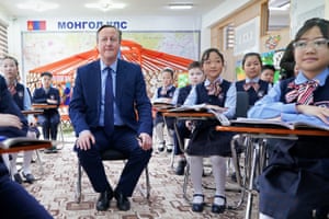 David Cameron sits on a small chair surrounded by children at their desks