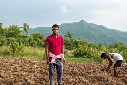 A man stands on ploughed ground next to another man bending down to the ground. There are trees and a green mountainside behind them