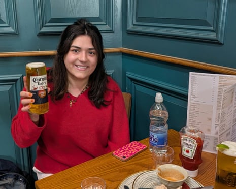 A woman in a red sweater holds up a glass of Corona beer while sitting at a restaurant table.
