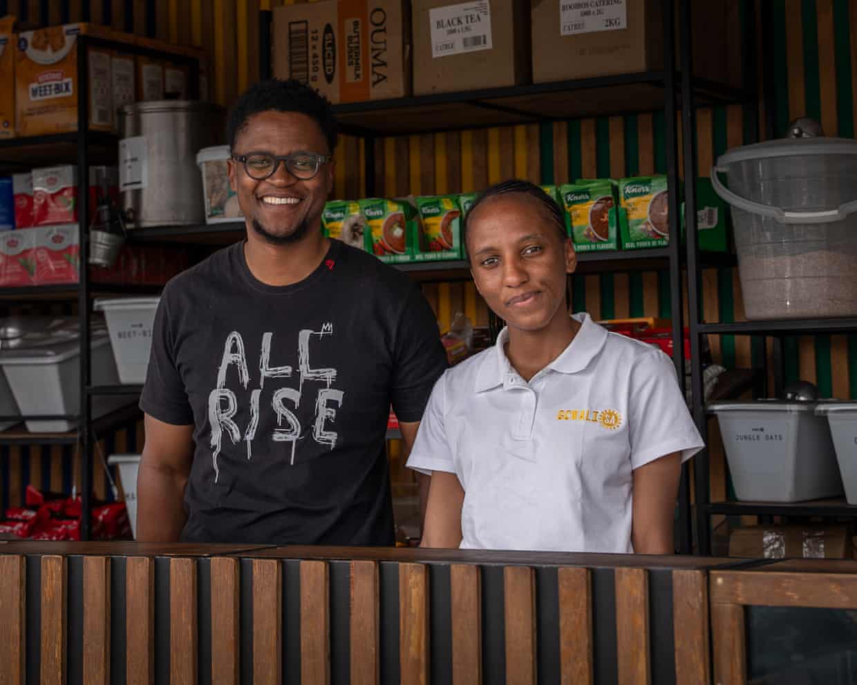 An African man and woman stand behind the counter in a small grocery shop in a shipping container