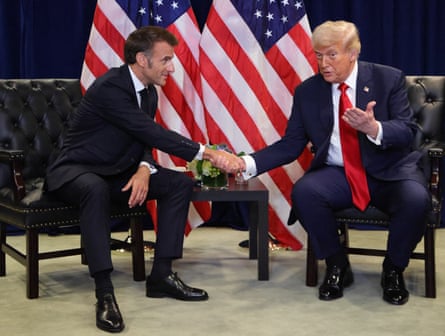 Macron and Trump shake hands; they are sitting on low chairs in front of two US flags. Trump is speaking and holding his other hand up and Macron is smiling as he listens and leans towards him.