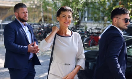 Sahra Wagenknecht getting out of a car, in a off-white dress, flanked by two security guards in blue blazers