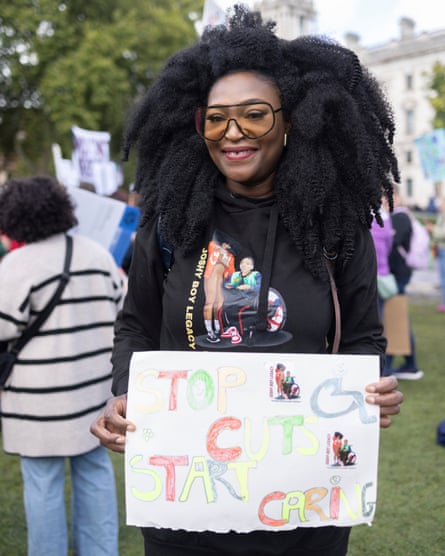 she holds a handwritten sign that reads 'stop cuts, start caring'. She wears a black hoodie with what appears to be an image of herself and her son, who uses a wheelchair, and the words 'Joshy boy legacy'; she wears yellow-tinted sunglasses and has long afro hair.