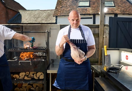 A man in a white T-shirt and a chef's apron, holding a leg of meat