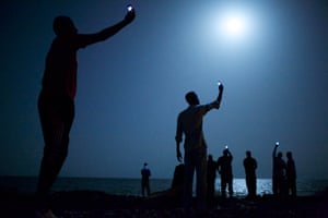 African migrants on the shore of Djibouti City at night under a full moon hold their phones aloft. The light of the screens is visible.