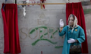 A woman wearing a mask of Queen Elisabeth stands next to the message by the elusive artist Banksy