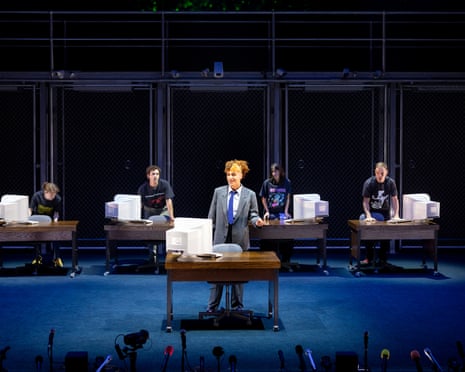 The actors stand at five desks, each bearing a large computer