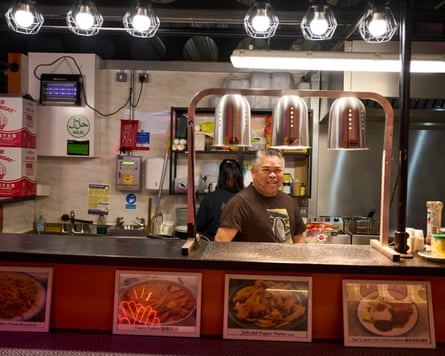 A man in a t-shirt stands in a small food stall smiling at the camera.