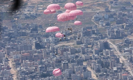 View from the open rear cargo door of a plane showing packages suspended on pink parachutes in midair over a devastated cityscape