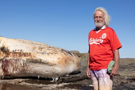 A man in a red football shirt stands next to the whale carcass