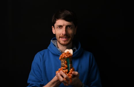 A man in a blue jumper, standing against a pitch-black background, holding an open tin of baked beans. The baked beans are spilling onto his hands. He looks concerned.