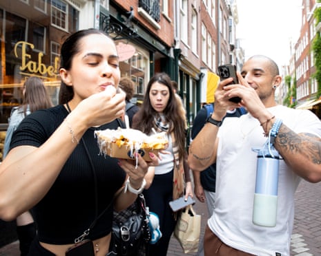 A man films a woman eating chips on the street outside a shop on his smartphone