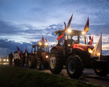 A convoy of tractors decorated with Serbian flags and accompanied by protesters on foot drives against a breaking dawn sky