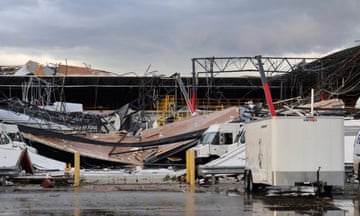 A large warehouse with the roof caved in, vehicles in the foreground, with wet pavement and a cloudy sky.