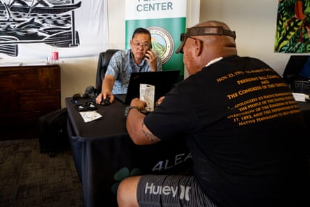 a man sits behind a desk on the phone using a computer as another man sits in front of him