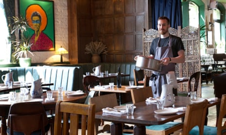The dining room at Parakeet, with wooden tables and pale blue seat-padded chairs, a pale blue banquette, and a chef in an apron carrying a large pot