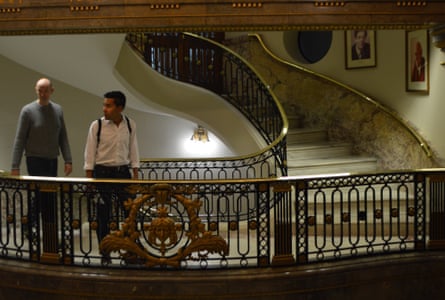 An ornate stairwell in the inside of Australia House. Two people pass by the landing and portraits hang on the wall.