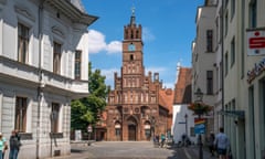 Brandenburg an der Havel, Germany: view down a wide street of historic buildings in the old town to the statue of the medieval knight Roland von Brandenburg outside the red-brick old town hall, a tall late Gothic building with arched doorways and windows and a tall, square clock tower.