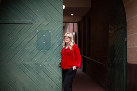Suzie Miller, wearing a red top and black pants, standing leaning on a green wooden door outdoors