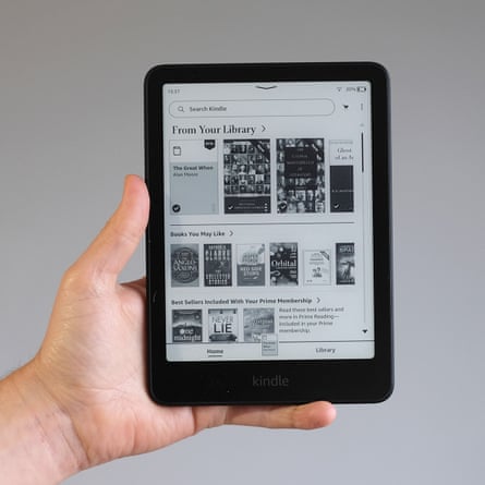 A hand holds a Kindle Paperwhite e-reader in front of a grey wall.