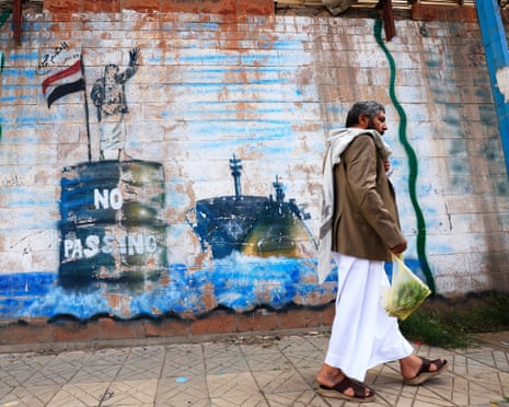 A man walks past a Houthi mural of a figure on a barrel labeled "No Passing" beside a ship