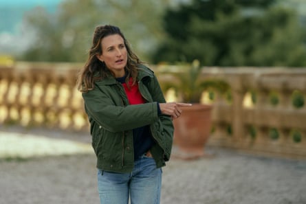 A woman in a green bomber jacket and jeans with long hair pulled off her face, walking on a sunny terrace, points off to the right of the image