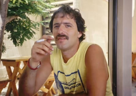 A man wearing with short brown curly hair and a moustache, wearing a yellow singlet and holding a cigarette
