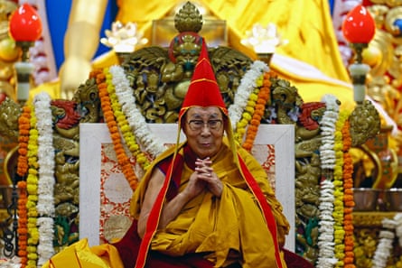 An elderly Buddhist monk in saffron robes sits on a throne decorated with flowers
