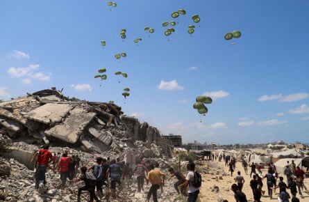 Palestinians run towards parachutes in the distance behind the rubble of destroyed buildings