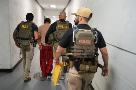 Men in tactical gear escort a man in handcuffs in a corridor.