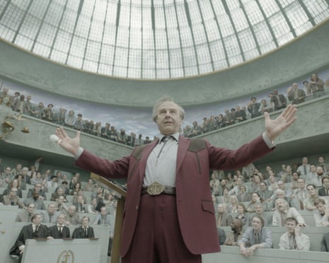 An ageing, overweight man standing arms outstreched in front of a circular lecture theatre full of people