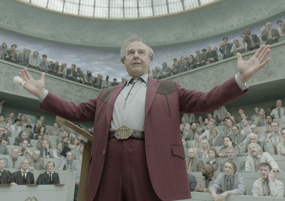 Tom Cruise (as Digger Rockwell) wearing a brown suit, stands arms open wide, appearing to address a packed hall