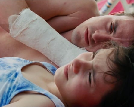 A shirtless man with a plaster cast on his upper arm lies on his side looking at a young girl in a vest