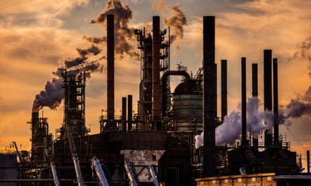 A sunset image of orange clouds, with dozens of smokestacks leaking smoke or steam into the air in silhouette.