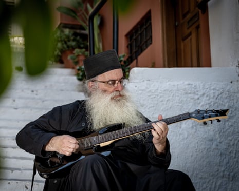 An Orthodox priest with a long white beard plays an electric guitar while sitting on whitewashed steps