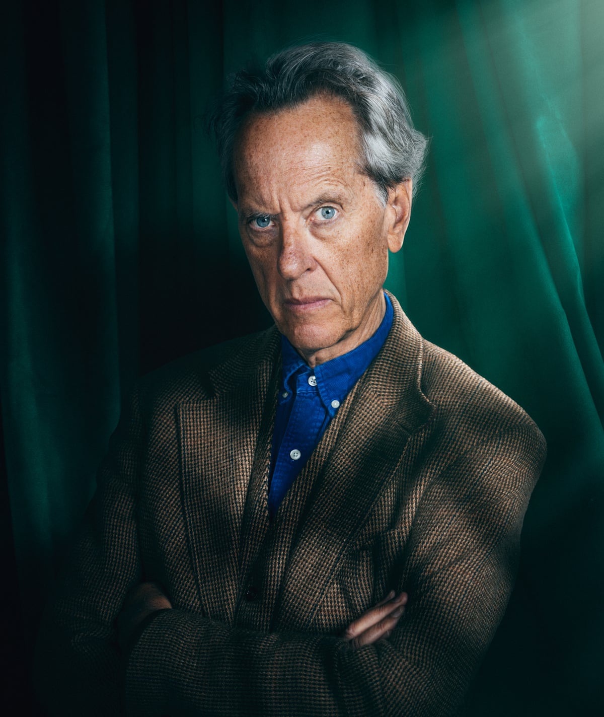 A man in a brown jacket, a blue shirt and short grey hair and blue eyes