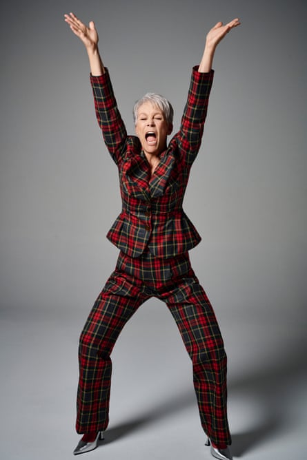 Portrait of Jamie Lee Curtis in a tartan suit and silver shoes crouching down with her hands raised high above her head and with her mouth open as if she is screaming