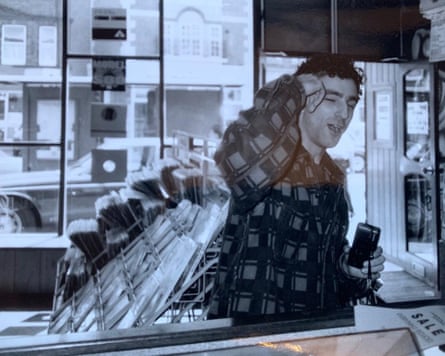 DJ Hype, with curly hair in a chequered shirt, laughs while holding a camera in a record shop
