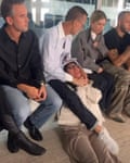 A man sitting at a fashion week presentation restrains a person on the ground by holding their neck with his hands and wrapping his foot around their body