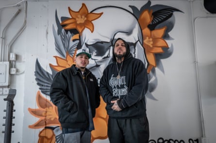 Two men stand in front of a wall with a painting of a skull and flowers on it.