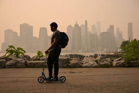 A man on a scooter rides through a tarmac area in a park with the Manhattan skyline behind him