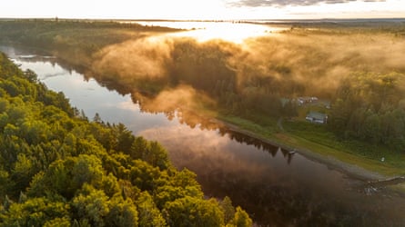 Mist rises above the trees on the banks of a sunlit river. There are a few buildings on one bank, in a clearing in the trees