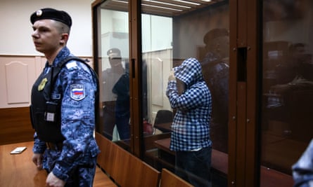 A man in a defendant's cage pulls a hoodie over his head while a guard stands watch