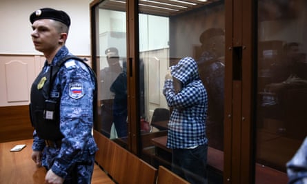 A man in a defendant’s cage pulls a hoodie over his head while a guard stands watch