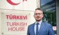 Yunus Paksoy standing outdoors in a suit next to a sign reading: 'Türkevi, Turkish House"