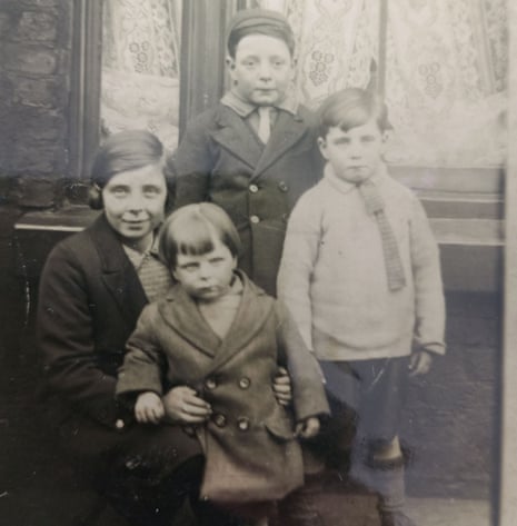 A faded black and white photograph of children in the 1940s. They are all wearing buttoned-up coats and one on the right is wearing a jumper