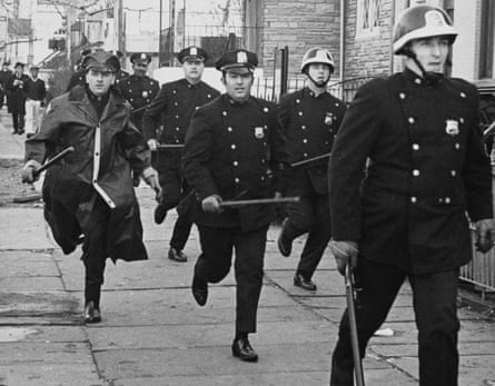 police, including some in helmets, hurry down a street in a black and white photo