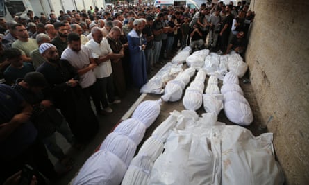 A crowd of men, heads bowed, mourn in front of bodies in bags