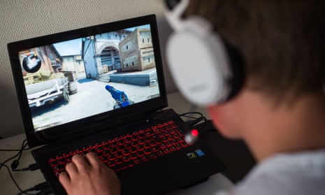 A young man wearing headphones playing a first-person shooter game on a laptop with illuminated skins